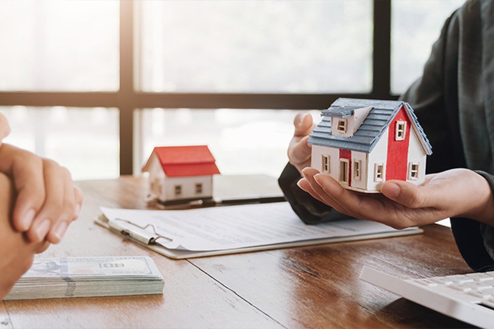 A person holding a miniature house with documents and money above the table