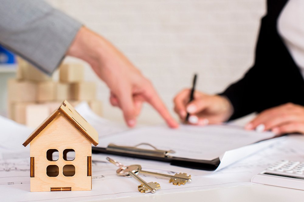 A wooden miniature house with keys beside it, and two persons signing of documents in the background