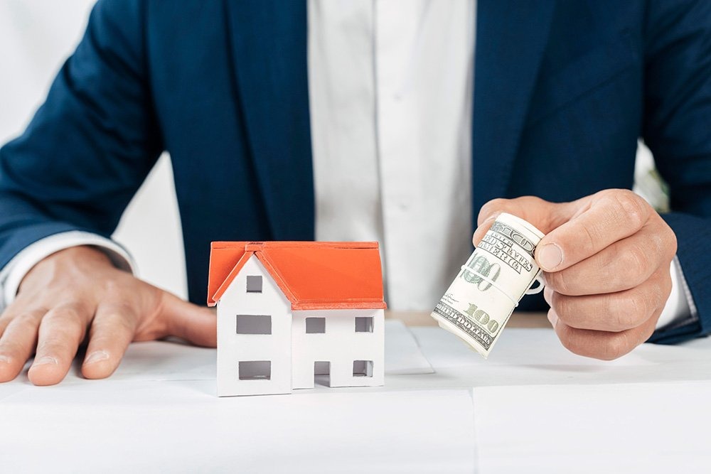 A man holding money in his left hand, and right hand placed above the desk with mini house