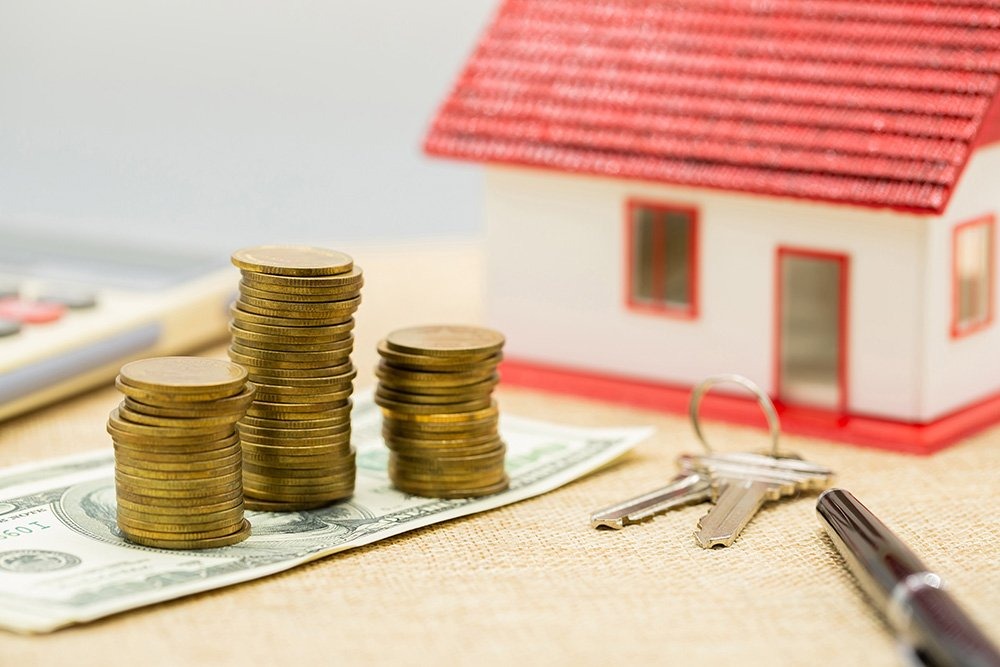 A dollar under some stack of coins, two keys and a pen beside, and calculator and mini house in the background