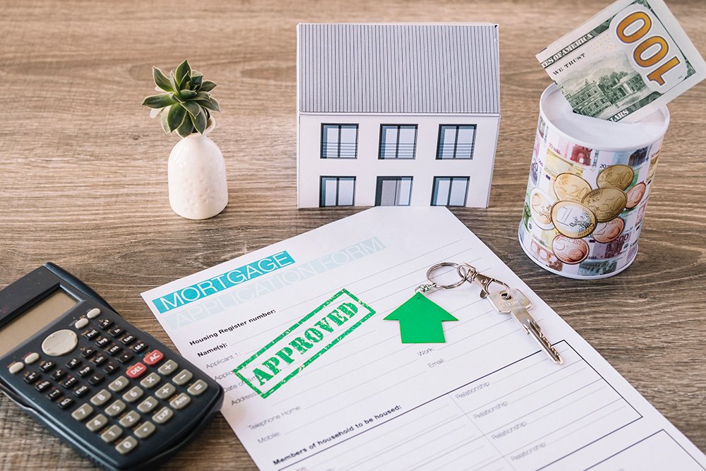 A calculator, miniature house, flow vase, money, key, and approved mortgage application form placed above the wooden table