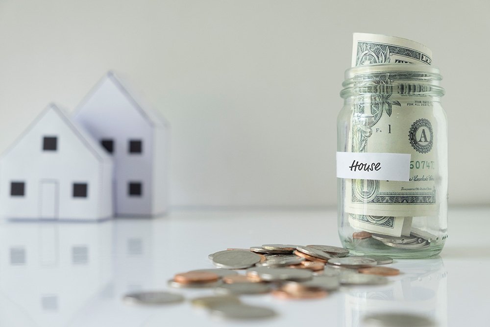 A glass jar labeled as house with money inside and some coins outside, and 2 mini houses in the background