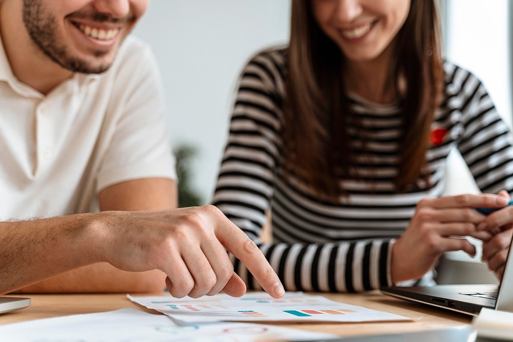 Two person smiling, a man pointing at the paper above the table